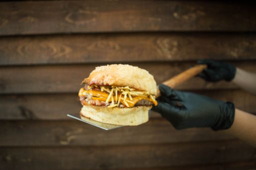 A close-up shot of a chef holding a juicy burger on a wooden board in the background of a wooden textured wall.