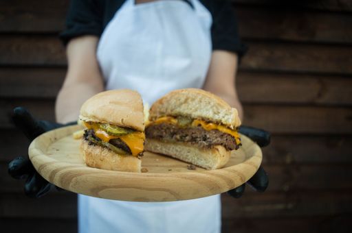 A close-up shot of a chef holding a burger cut in half on a wooden plate.
