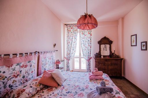 A brown dresser with mirror in a pink room with curtained windows near flower patterned bed with pillows