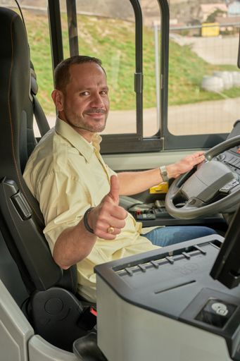 A Hispanic male driving a modern bus in the daylight with a garden outside