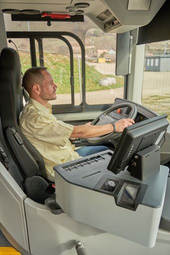 A Hispanic male driving a modern bus in the daylight with a garden outside