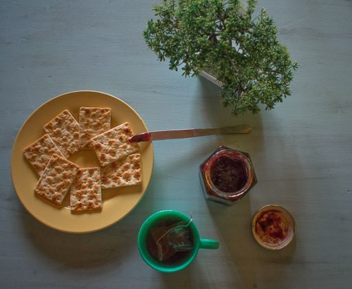 A high angle view of crackers on a plate on the table with a cup of tea and jam under the lights
