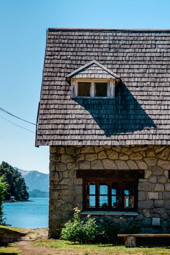 A vertical shot of a stone house with a dormer window near the scenic lake and mountains