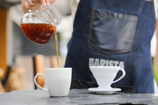 A barista pouring a coffee from a glassy pot into a cup on the table