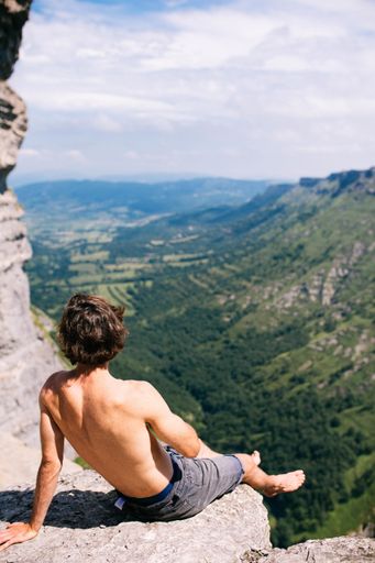 A vertical shot of a lonely male sitting on top of a rocky cliff and enjoying the beautiful view of mountains and greenery