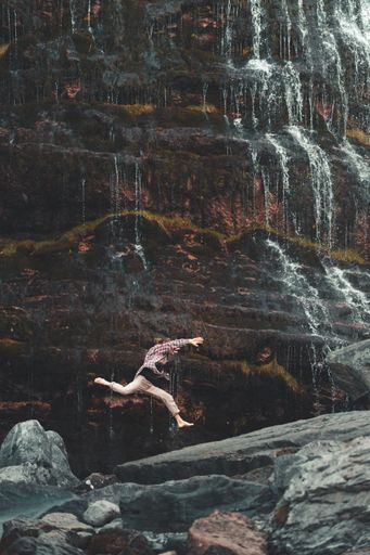 A vertical shot of a person jumping on a rock near a cascade during a sunny day-concept of freedom