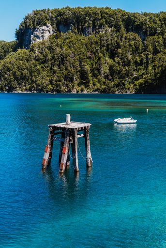 A vertical shot of a wooden mooring dolphin in turquoise water surrounded by mountains on a sunny day
