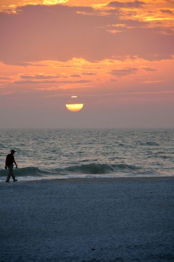A vertical shot of a person walking on the beach with the sunset sky in the background