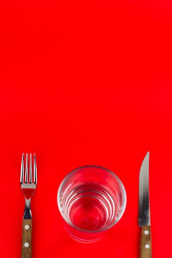 A vertical shot of an empty glass with a fork and knife beside it on red background