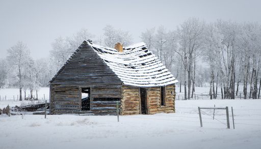 A view of an old abandoned cabin house in the field covered with snow in the background of leafless trees.