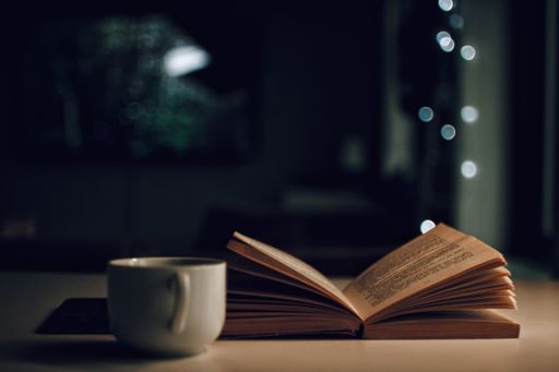A closeup of an opened book next to a ceramic coffee cup on the table with dark blurred background