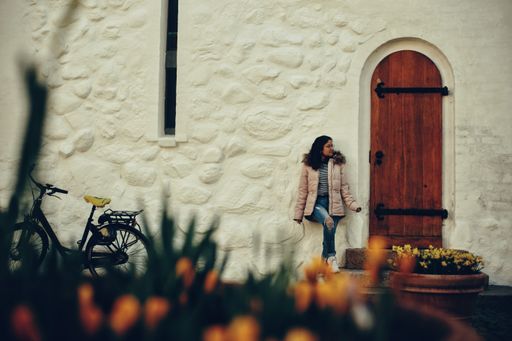 A beautiful shot of a South Asian girl standing with her back leaning against white stone wall beside an old wooden door during daytime