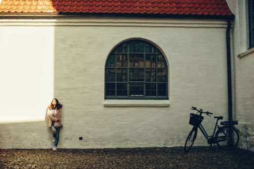 A beautiful shot of a South Asian girl standing with her back leaning against white stone wall near to a big window with parked bicycle close to her