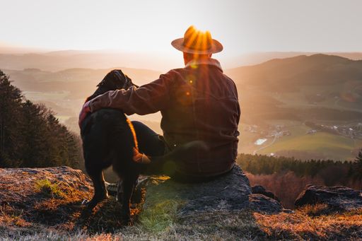 A man with a dog enjoying a beautiful view while sitting on top of a rocky mountain during sunset