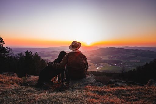 A man with a dog enjoying a beautiful view while sitting on top of a rocky mountain during sunset