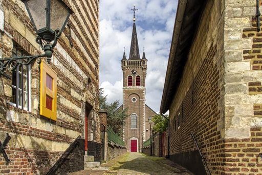 A narrow street between brown houses leading to the high building with a cross on top  under the cloudy sky