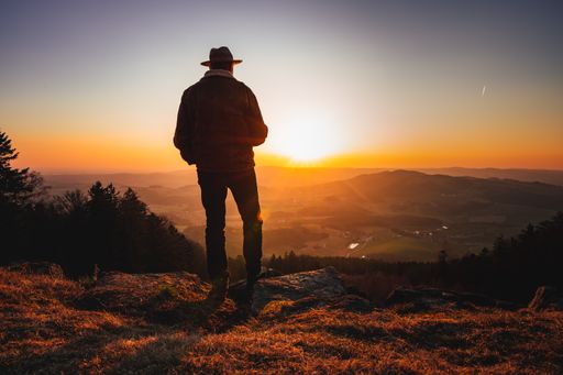 A man with a hat standing on a mountain peak and enjoying a beautiful landscape during sunset