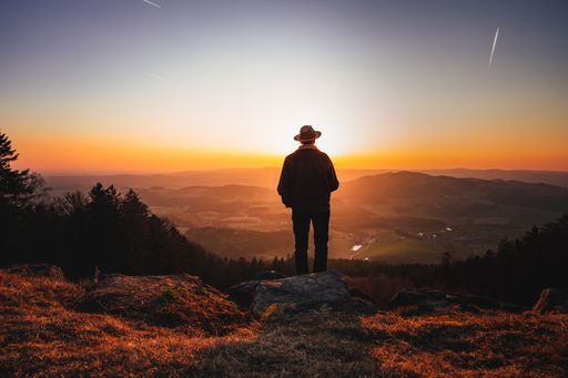 A man with a hat standing on a mountain peak and enjoying a beautiful landscape during sunset