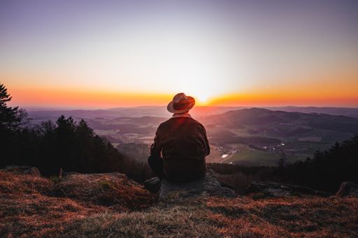 A rear view of a man with a hat enjoying a beautiful view while sitting on top of a rocky mountain