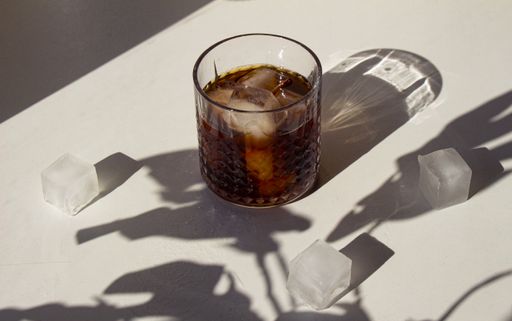 A closeup of a glass of coffee with ice cubes inside and next to it on a white table in the sunlight