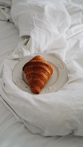 A vertical shot of a croissant on a white plate on a white fabric surface. Breakfast in bed concept