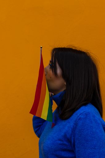 A vertical portrait of a woman kissing a pride flag against a yellow background