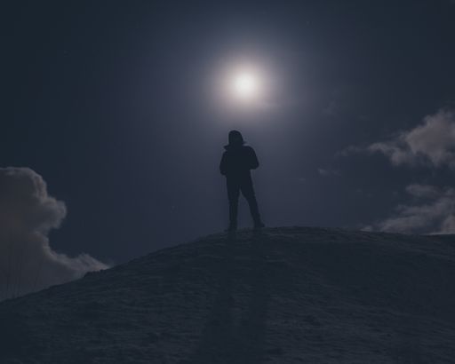 A silhouette shot of a male standing on a hill under moonlight in the night sky with clouds