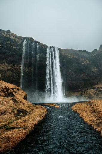 A vertical shot of a river in the background of a waterfall coming out of a cliff.