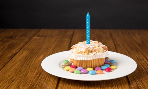 A plate with muffin with burning candle and colorful candies on a wooden table near gray wall
