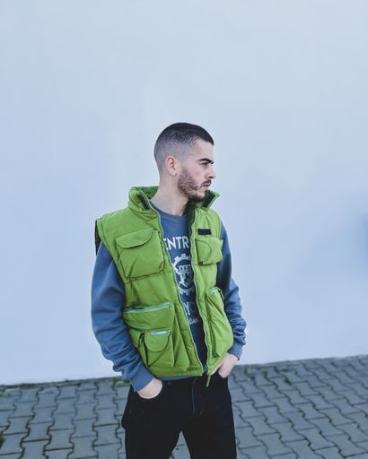 A vertical shot of a young man wearing green gilet and black pants posing next to a white wall