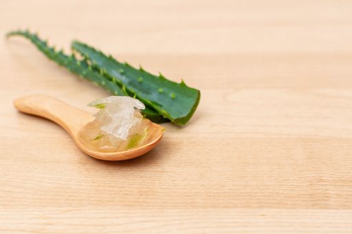 A close-up shot of Aloe-Vera gel in a wooden spoon with Aloe-Vera green peels on a wooden table with blurred background