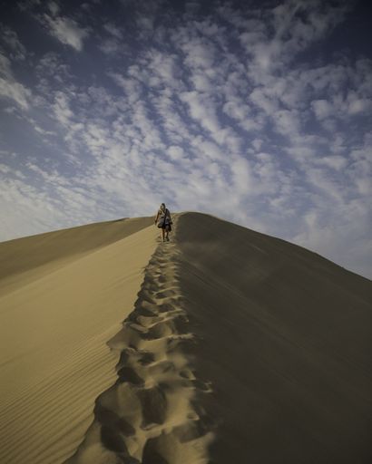 A vertical shot of a person walking through sand dunes under a cloudy blue sky