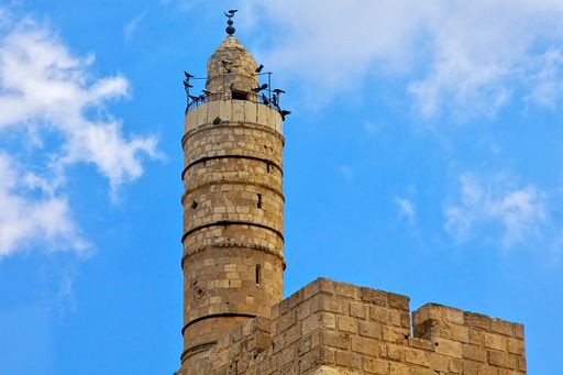A low angle shot of the Tower of David in Jerusalem with birds on the top and a clear blue sky