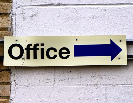 A closeup of a metal Office sign with a blue arrow and a crack on the white brick wall background