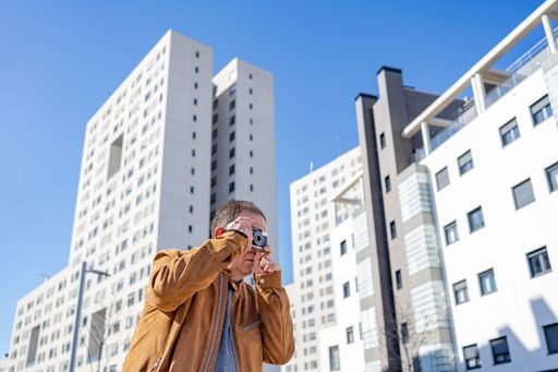 A older man photographing with an old film camera in the city with buildings in the background.
