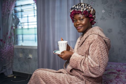 Beautiful Black girl wearing satin bonnet and morning coat, holding a cup and side plate. smiling and looking at camera - concept on Africa millennial