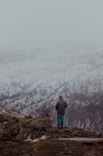 A vertical shot of a man standing on a cliff enjoying the beautiful snowy landscape with a toy car on the ground