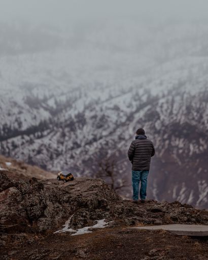 A back shot of a young man with his toy car on the mountain top against snowy mountain forest during daytime