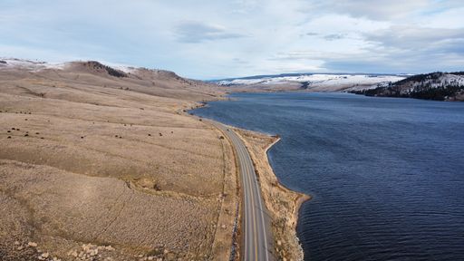 An aerial view of an asphalt road going by a lake and rocky hills with snowy mountains on the horizon on a sunny day