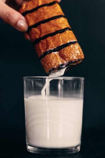 A vertical image of a person dipping a chocolate glazed pastry into a glass of milk against a black background