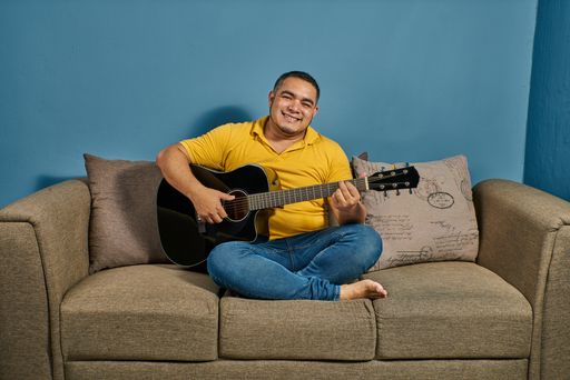 A Hispanic man on a sofa in his house practicing guitar