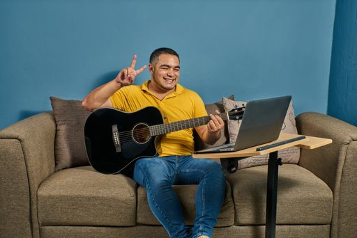 A Hispanic man on a sofa in front of his laptop Greeting fellow students in the online guitar class