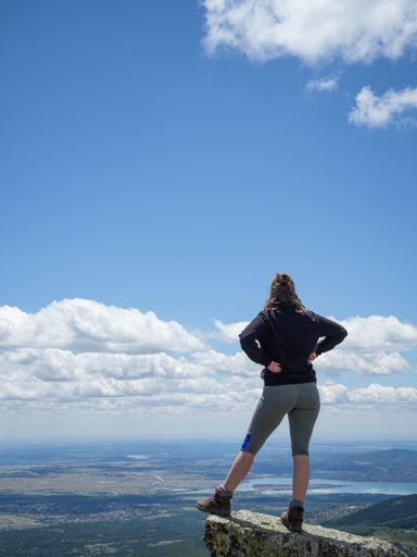 A vertical shot of a female standing on top of a cliff with an overlooking view of fields, mountains, and river