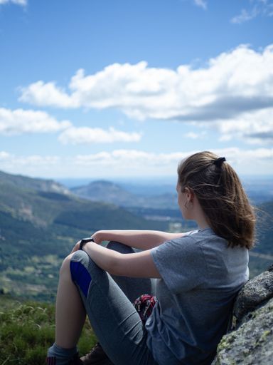A vertical shot of a female sitting on a grass surface with a beautiful view of mountains under a cloudy blue sky
