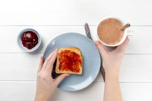 A top view shot of a hand holding toast with strawberry jam on a blue plate and cup of cacao on a white wooden background