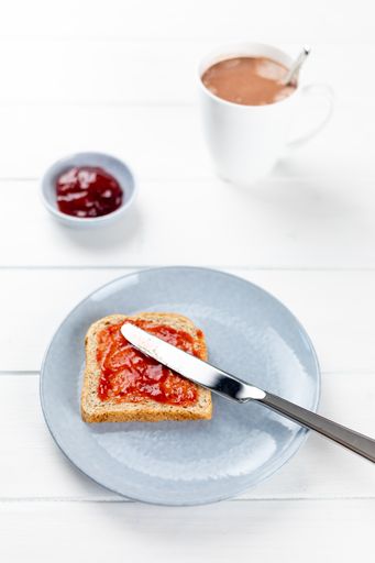 A closeup shot of toast with strawberry jam on a blue plate and a cup of cacao on a white wooden background