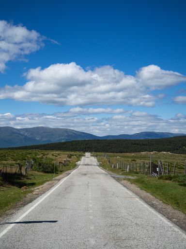 A vertical shot of a long road between green fields with mountains and a cloudy blue sky in the background