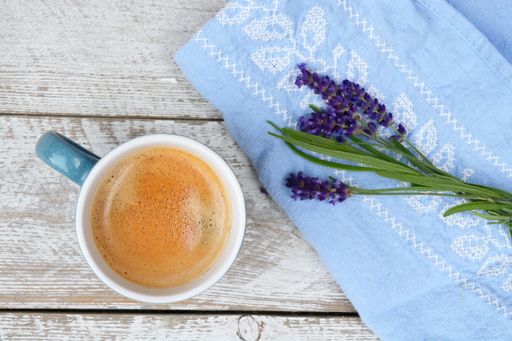 A closeup shot of a cup of coffee with lavender flowers and blue fabric on a wooden surface