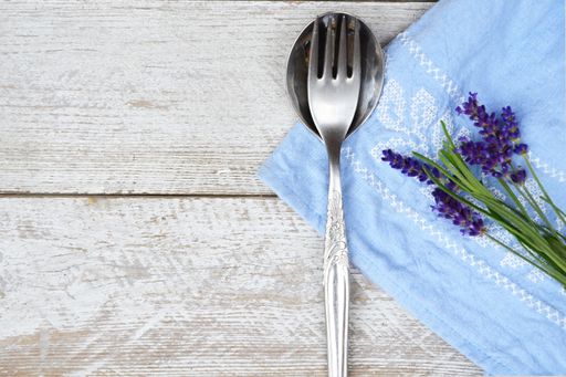 A closeup shot of a silver fork, spoon, and lavender flowers with blue fabric on a wooden surface
