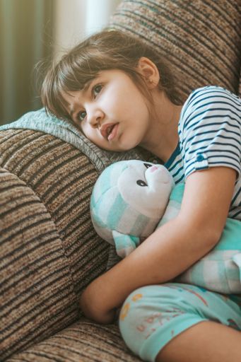 A vertical portrait of a little girl lying on the couch with a toy in her hands and watching a TV